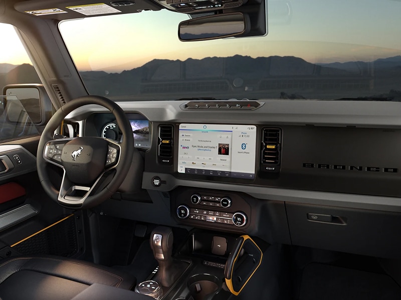 Ford Bronco interior with touchscreen and steering wheel at sunset, highlighting dashboard features.