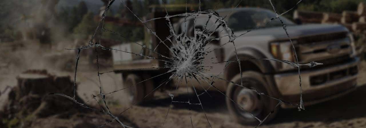 Cracked windshield view of a truck driving on a dusty road with trees in the background.
