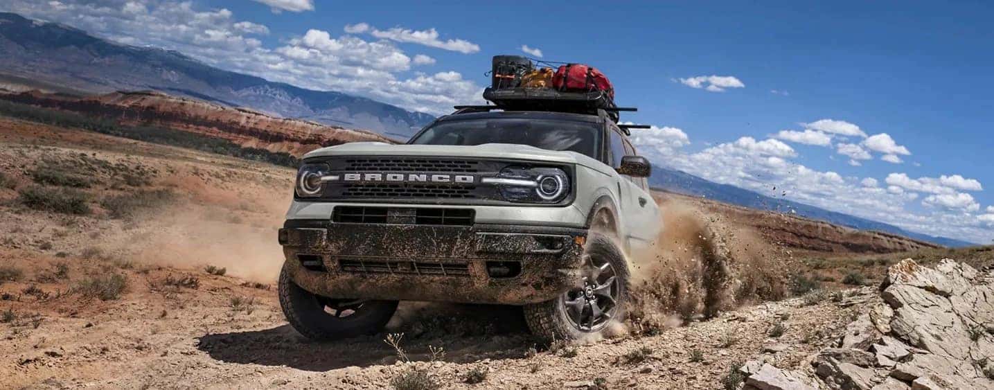 Off-road SUV driving through a muddy desert terrain, carrying gear on its roof, under a clear blue sky.