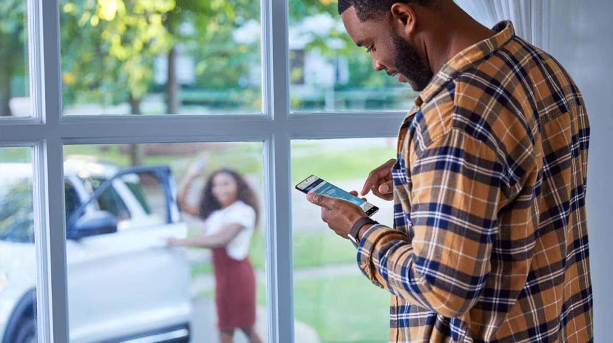 Man using smartphone by window, controlling car remotely. Outdoors, a woman waves near a white vehicle.