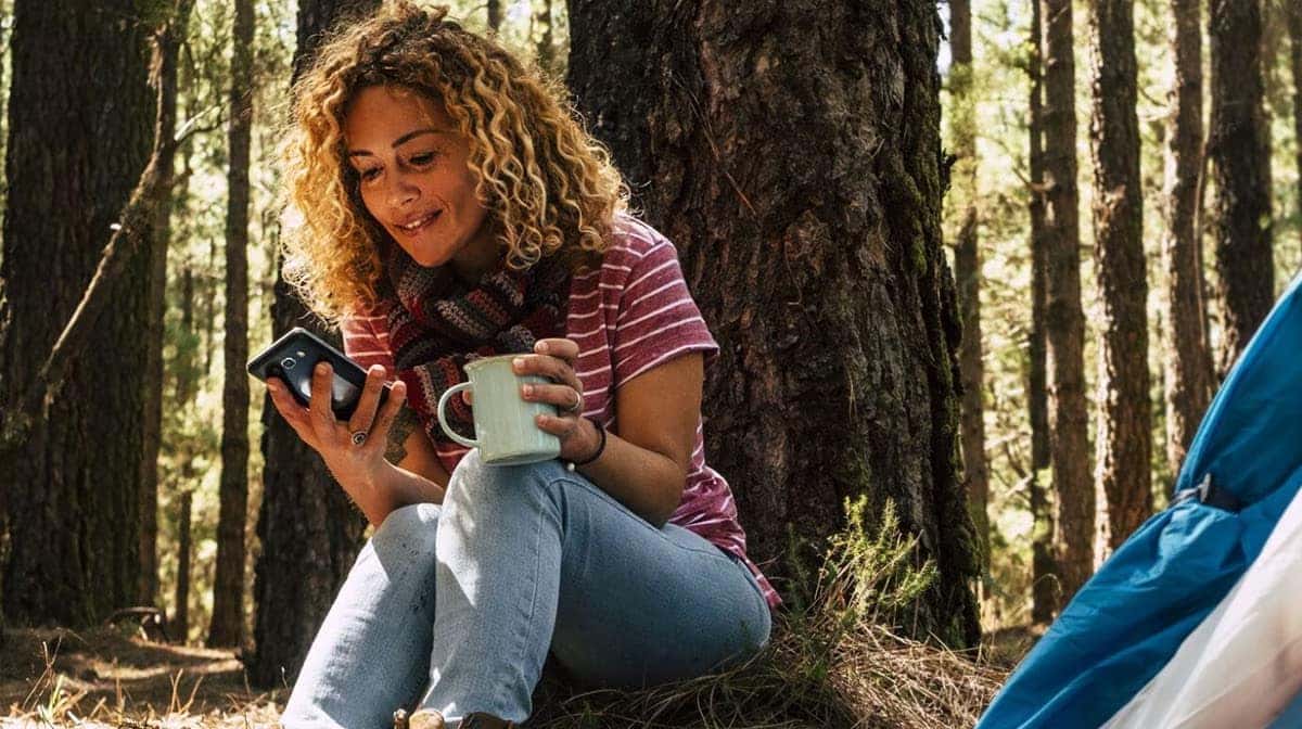 Woman with curly hair enjoys coffee in a forest, using her phone near a tent.