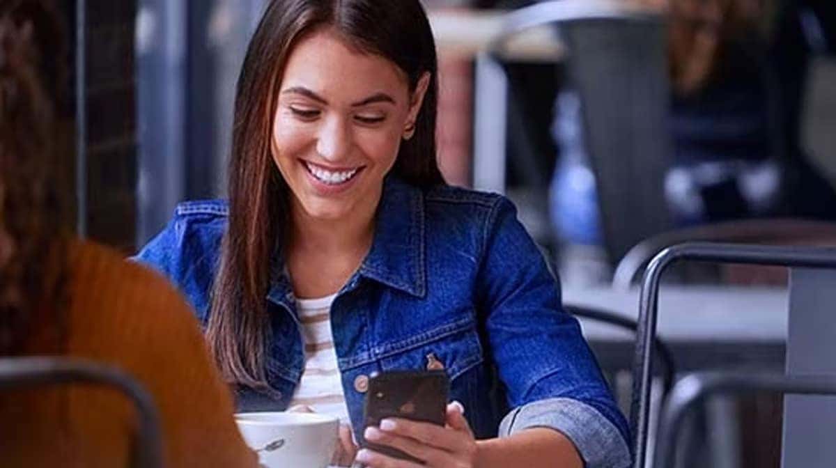 A woman in a café smiles at her phone, wearing a denim jacket and holding a coffee cup.