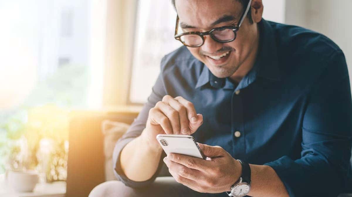 Man in glasses smiling while using a smartphone, wearing a blue shirt in a bright room.