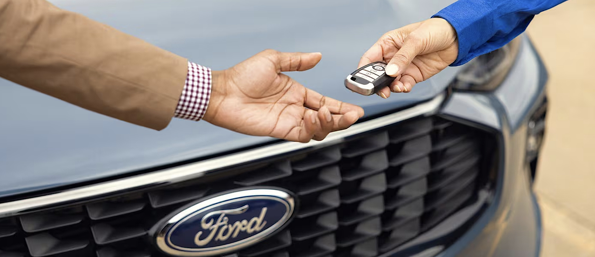 Hands exchanging a car key fob in front of a Ford vehicle grille, symbolizing car ownership or rental.