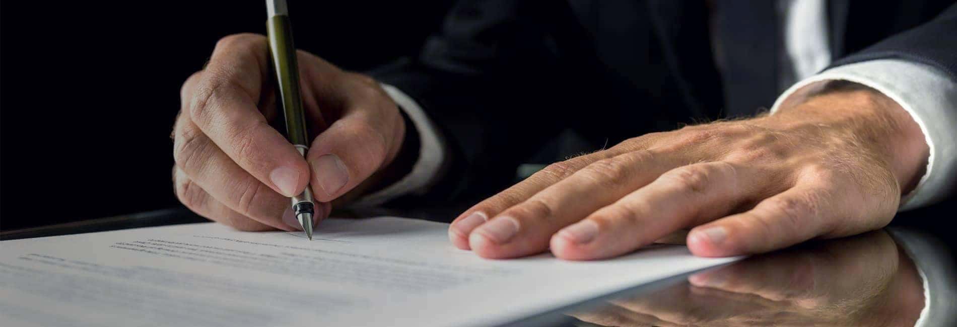 Person in a suit signing a document with a pen on a reflective surface.