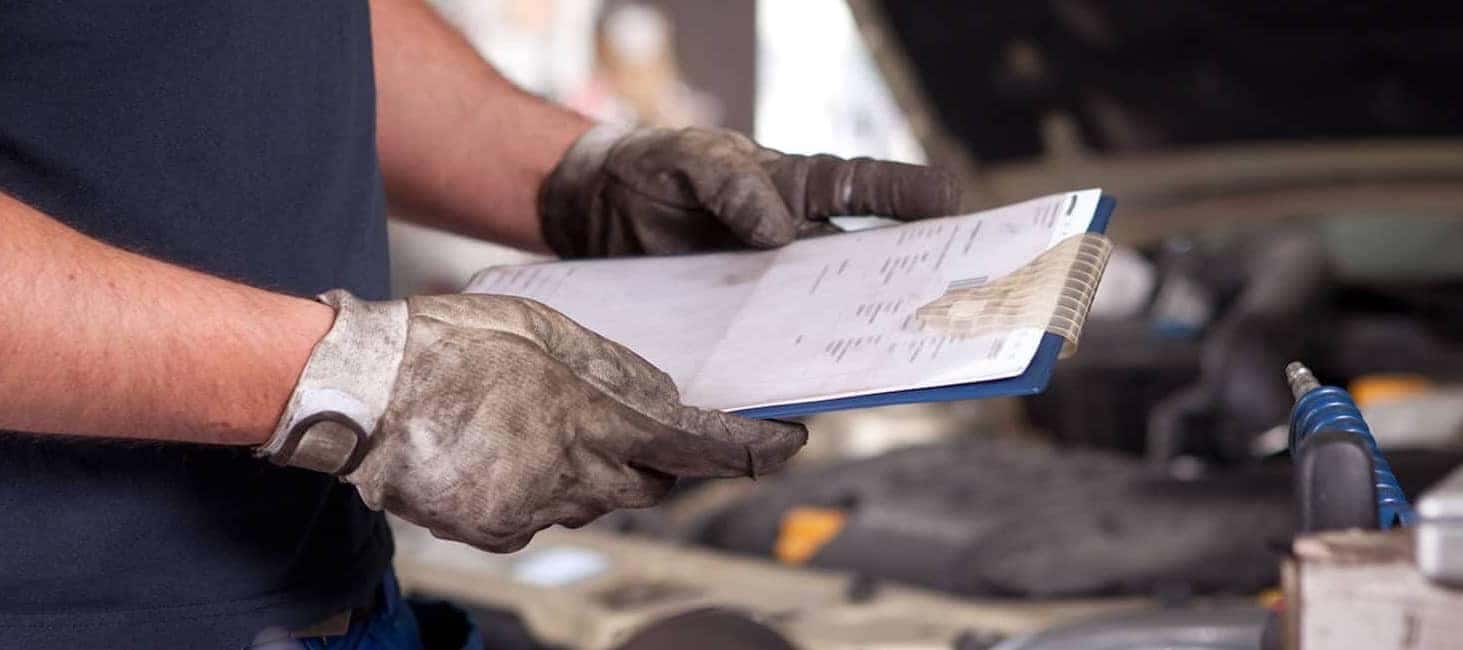 Mechanic with dirty gloves reviewing a clipboard near a car engine in a repair shop.
