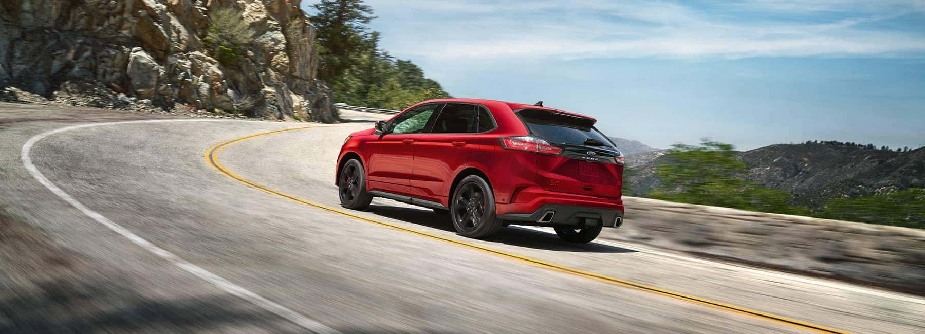 Red SUV driving on a winding mountain road with scenic rocky and tree-lined surroundings.