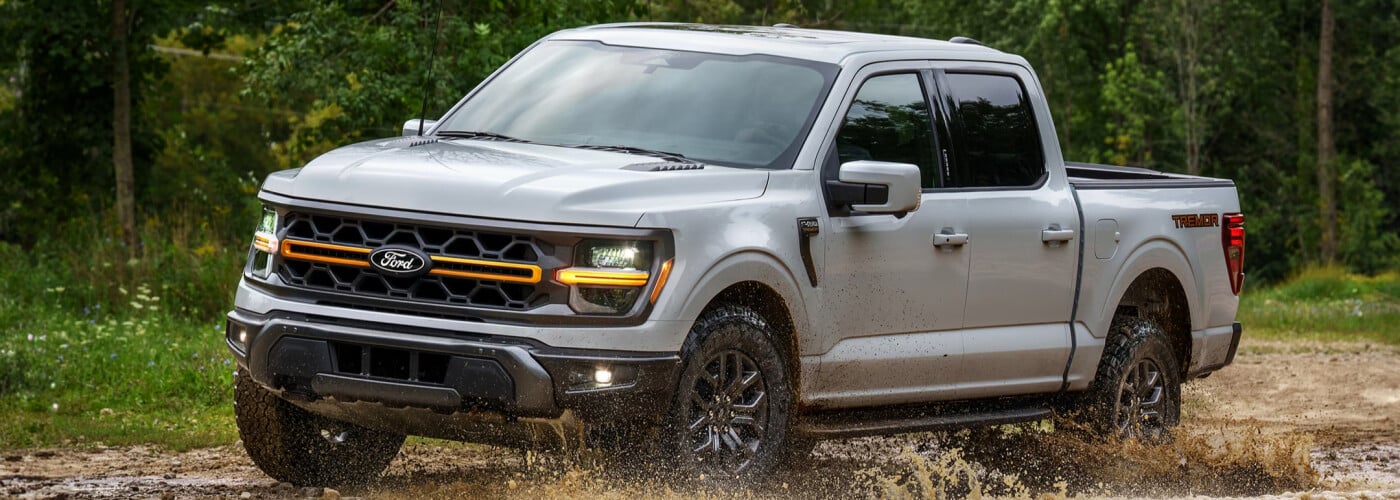 Silver Ford pickup truck driving through a muddy path in a forested area, showcasing off-road capabilities.