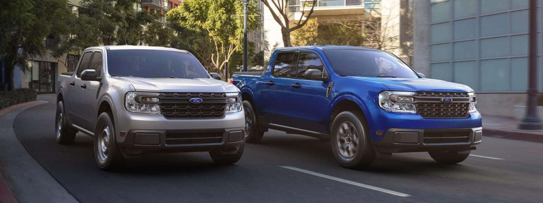 Two Ford trucks, one silver and one blue, drive on a tree-lined urban street.