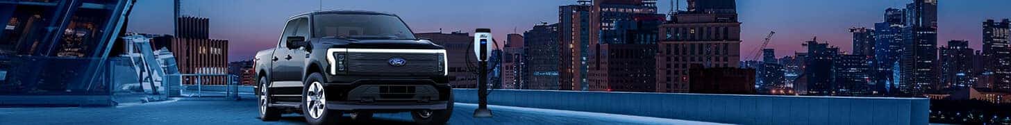 Electric truck charging at a charging station on a rooftop with a cityscape at dusk.