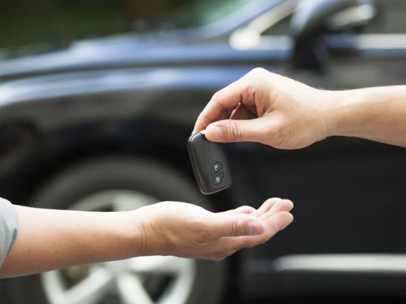 Hands exchanging a car key fob with a parked car in the background.